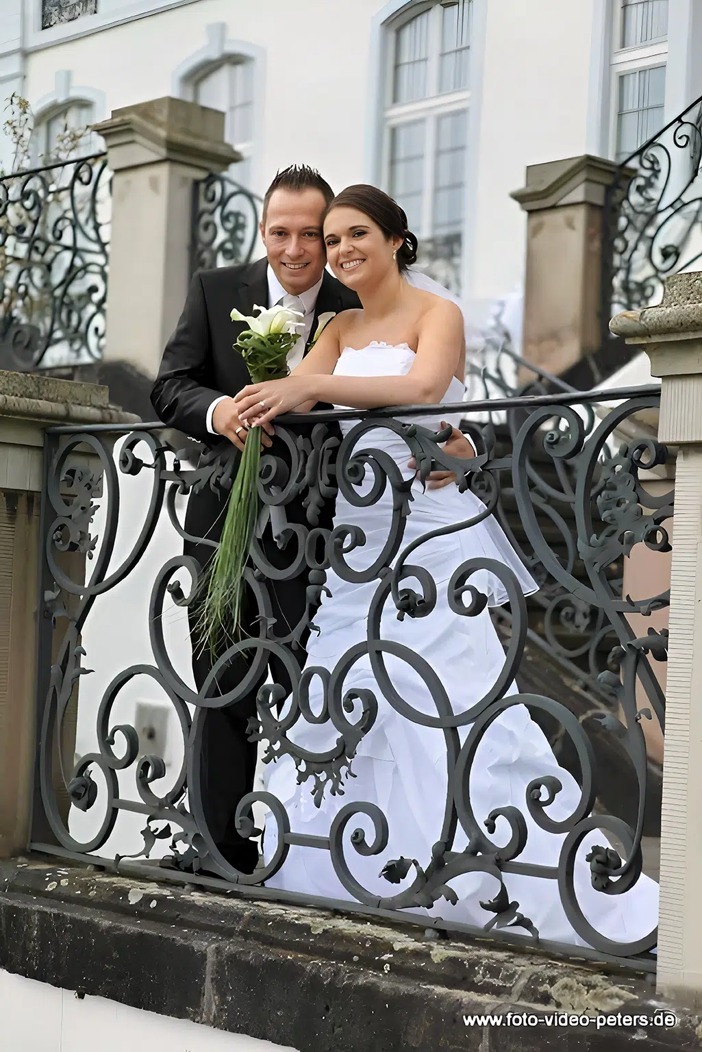 Hochzeitspaar steht eng aneinander gelehnt, während es sich kopf an kopf vor einem Geländer positioniert und in die Kamera schaut.