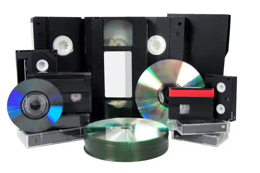 Eine Sammlung verschiedener analoger Medien liegt auf einem weißen Hintergrund. Zu sehen sind mehrere VHS-Kassetten, eine MiniDV-Kassette, eine Hi8-Kassette, eine Audio-Kassette sowie mehrere CDs und DVDs in ihren Hüllen und als Stapel.