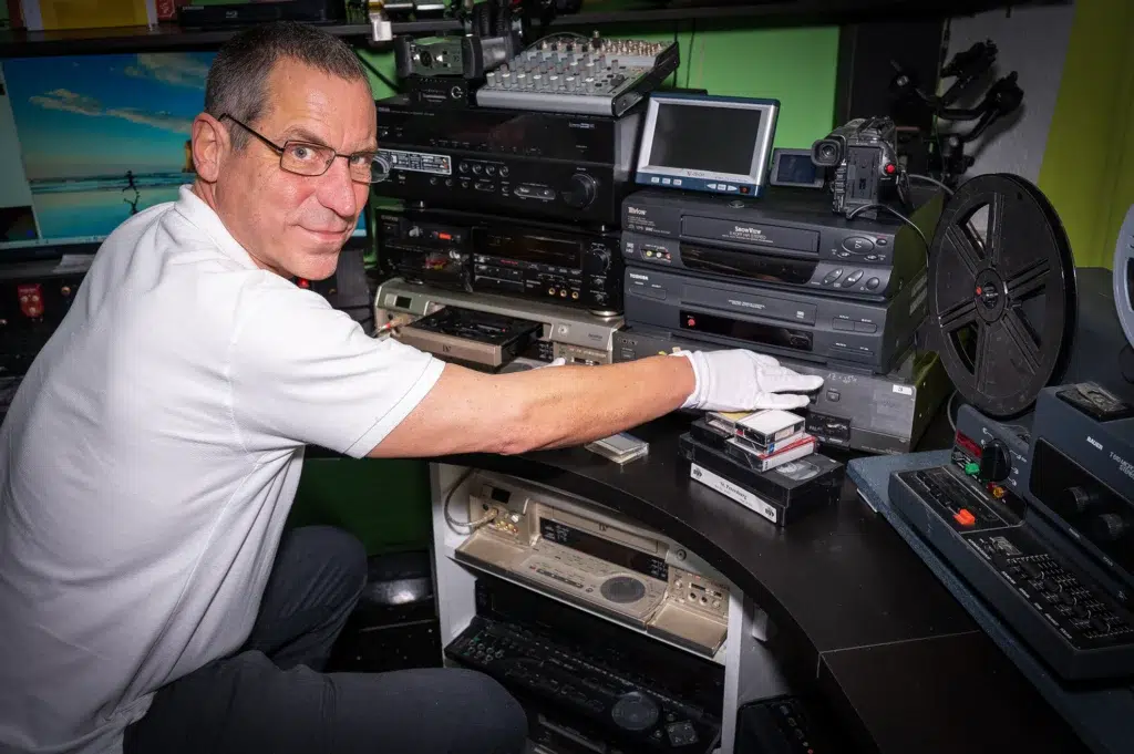 Ein Mann mit Brille und weißem Poloshirt kniet vor einem Schreibtisch voller Geräte zur Digitalisierung von analogen Medien. Er trägt weiße Handschuhe und hantiert mit einer kleinen Videokassette (vermutlich MiniDV). Hinter ihm befinden sich mehrere Videorekorder, ein Mischpult, ein kleiner Monitor und andere Digitalisierungskomponenten. Im Hintergrund ist ein Computermonitor mit einem Strandbild zu sehen.