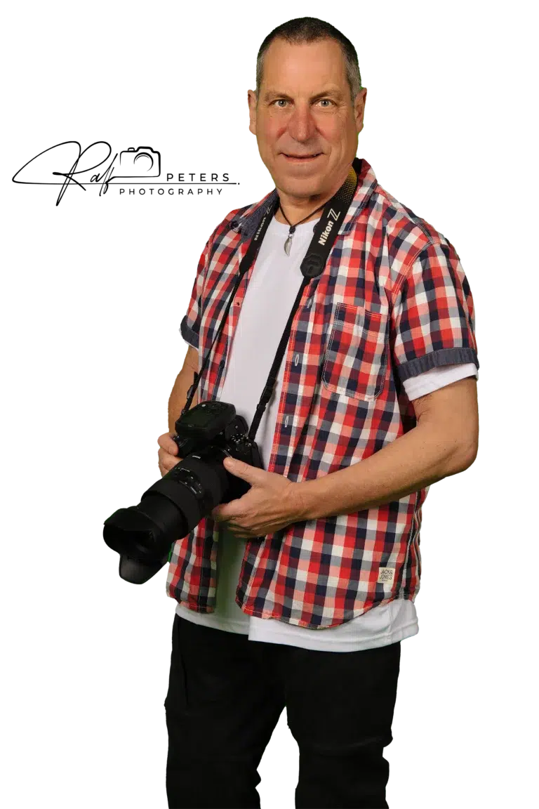 Ralf Peters, Inhaber von Foto-Video-Peters, Fotograf aus Boppard, hält professionelle Kamera. Er trägt ein kariertes Hemd über einem weißen T-Shirt und eine Kameragurt mit "Nikon Z" Aufschrift. Freundliches Lächeln.