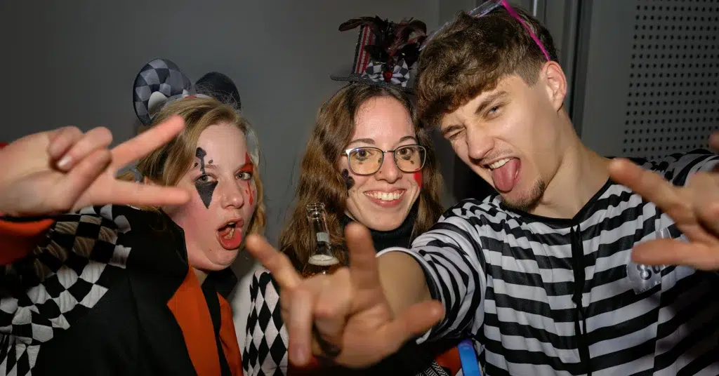 Drei kostümierte junge Leute (Harlekin, Harry-Potter-inspiriert, Gestreift) mit lustigen Gesten auf einer Karnevalsparty .