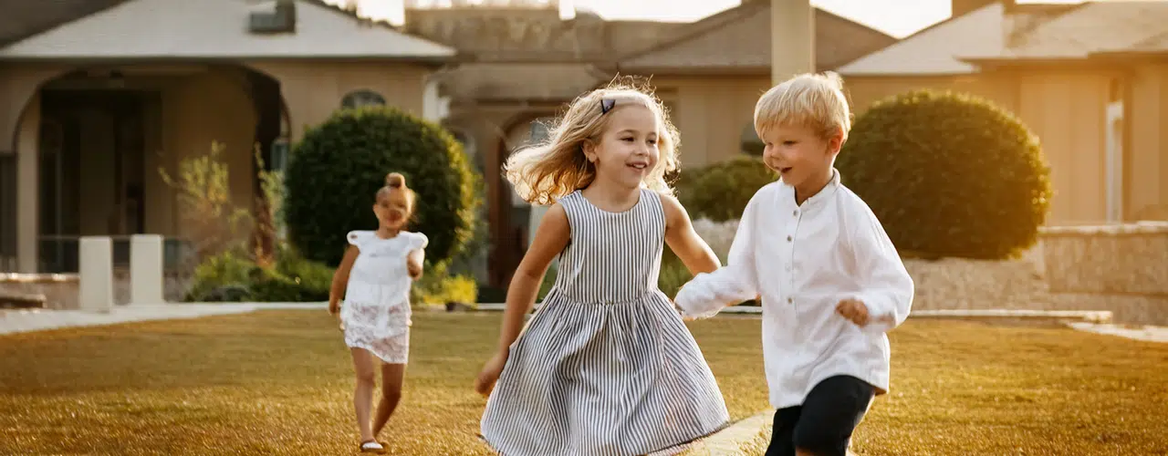 Ein sonniges Farbfoto zeigt drei fröhliche Kinder, die auf einem gepflegten Rasen vor einem Wohnhaus spielen. Ein blondes Mädchen in einem gestreiften Kleid hält Händchen mit einem blonden Jungen in einem weißen Hemd und dunklen Shorts, beide rennen lachend auf den Betrachter zu. Ein drittes Mädchen in einem weißen Kleid läuft etwas weiter hinten hinterher.