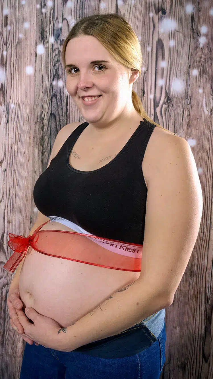Schwangere Frau lächelt in die Kamera, hält ihren Babybauch mit roter Schleife.