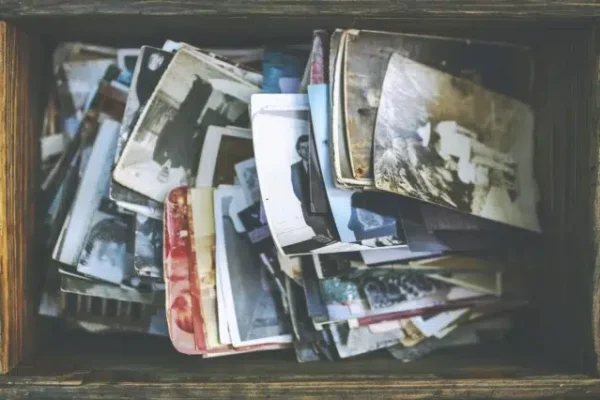 Ein überfüllter Holzkasten enthält einen unsortierten Stapel alter Fotoabzüge verschiedener Größen und in unterschiedlichem Zustand. Einige Fotos sind schwarz-weiß, andere farbig. Die Motive sind nicht klar erkennbar.
