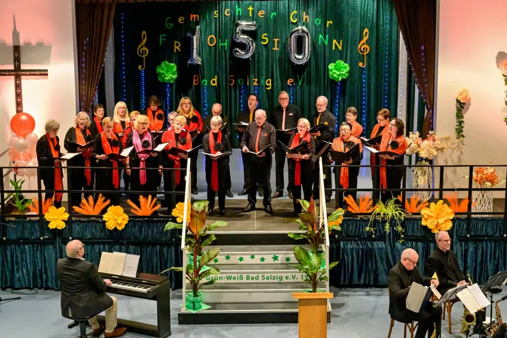 Gemischter Chor Frohsinn Bad Salzig singt auf der Bühne bei der Auftaktveranstaltung, begleitet von Klavier und Bläsern, mit festlicher Dekoration.