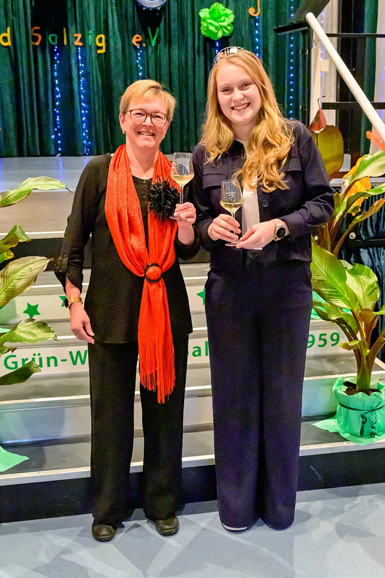 Zwei Frauen mit Sektgläsern lächeln in die Kamera bei einer Feier auf einer Bühne mit grüner Dekoration.