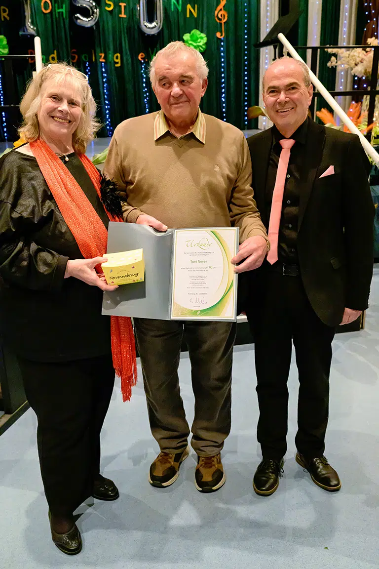 Drei Personen bei einer Ehrung auf einer Bühne, eine Frau hält eine Urkunde und ein Geschenk in der Hand.