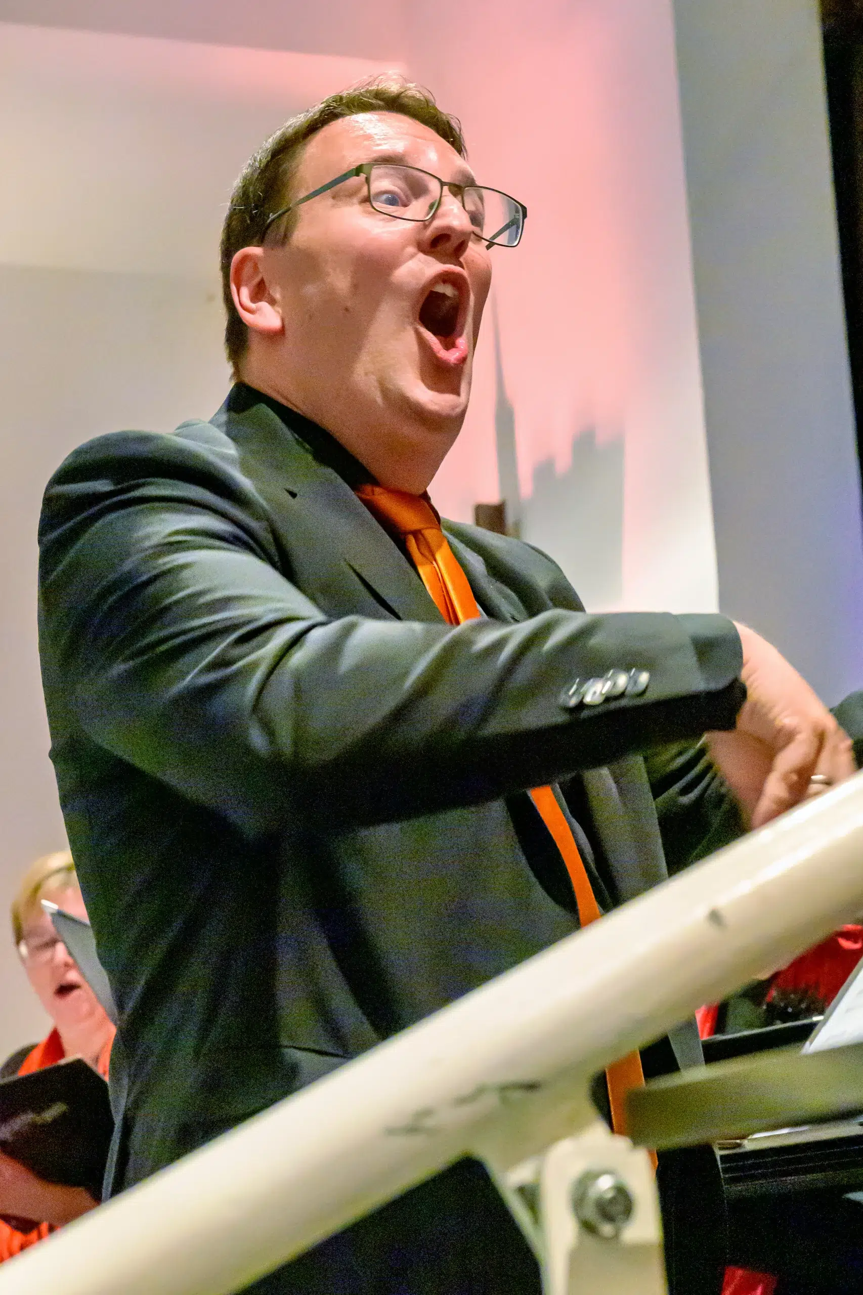 Dirigent mit Brille und orangener Krawatte dirigiert Chor bei einer Veranstaltung in Innenraumbeleuchtung.