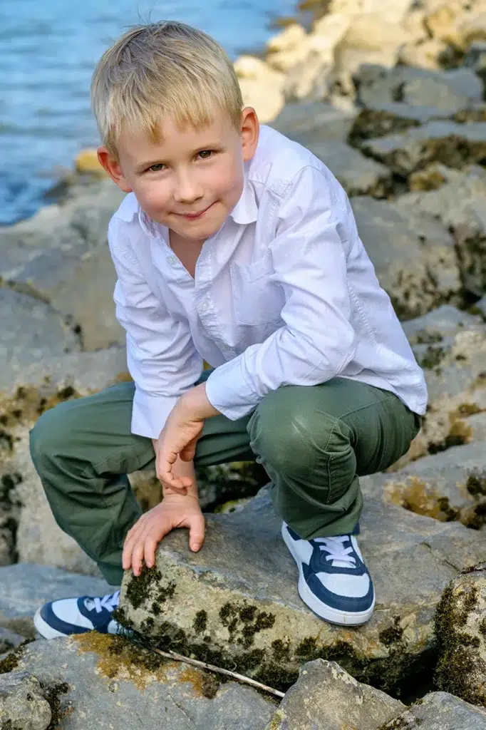 Ein charmanter, blonder Junge in einem weißen Hemd hockt auf den Felsen am Rheinufer