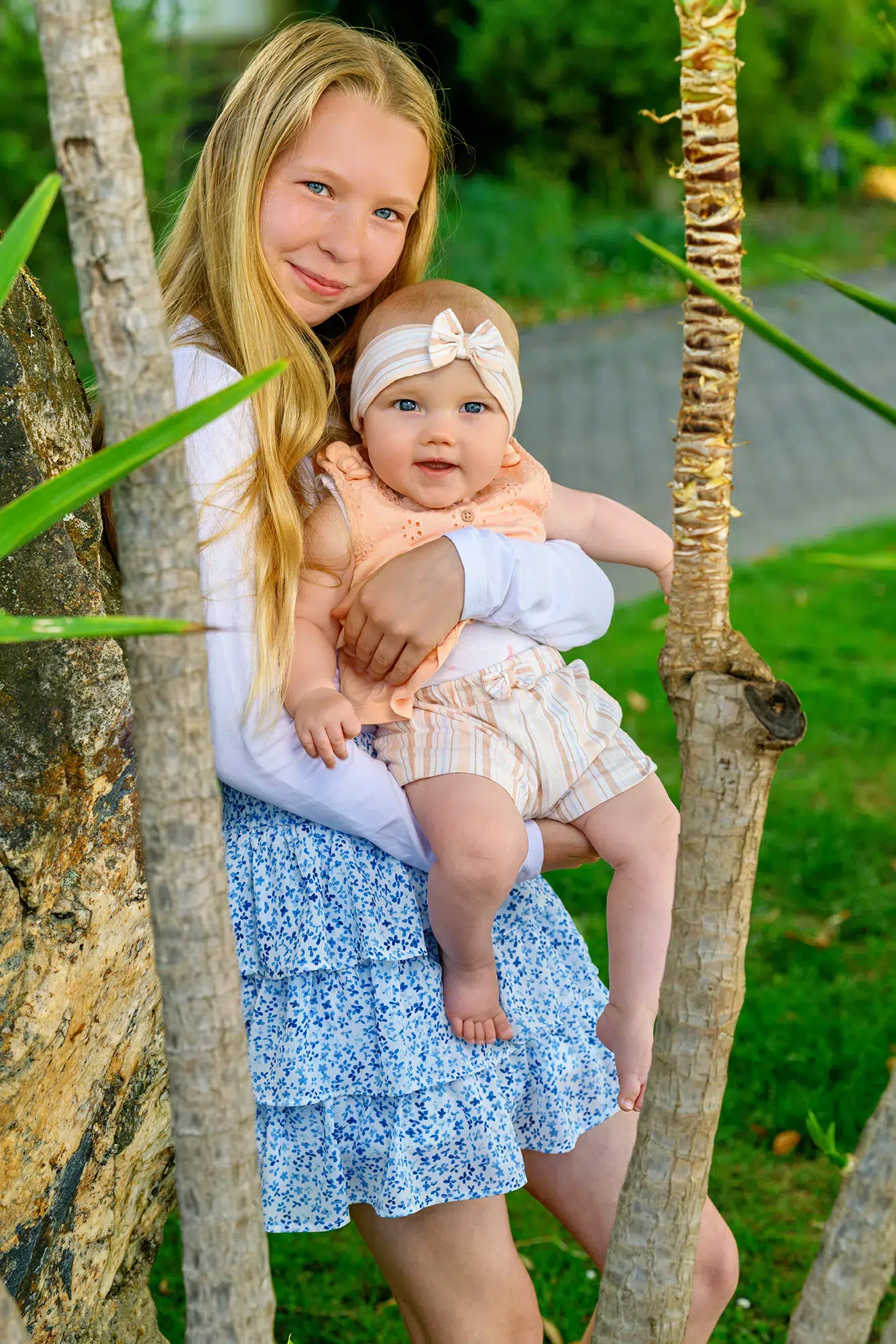 Eine stolze, ältere Schwester mit langen blonden Haaren hält ihr Baby-Schwesterchen liebevoll im Arm
