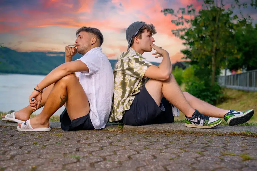 Zwei junge Männer sitzen Rücken an Rücken vor einem farbenfrohen Sonnenuntergang am Rhein,