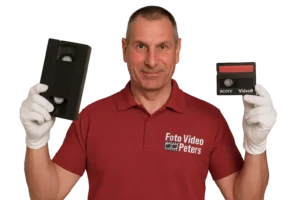 Mann im roten Polohemd mit Logo „Foto Video Peters“, weiße Handschuhe, hält eine VHS-Kassette und eine Video8-Kassette